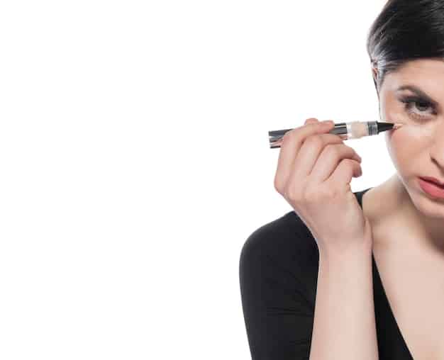 A woman applying black liquid eyeliner to her upper eyelid with a steady hand, creating a precise wing. The background is a well-lit vanity with various makeup products.