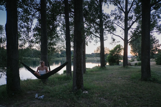 A person relaxing in a hammock strung between two trees in a peaceful forest setting. Sunlight filters through the leaves, creating a dappled effect. The person is holding a book and appears to be fully immersed in reading and relaxation.